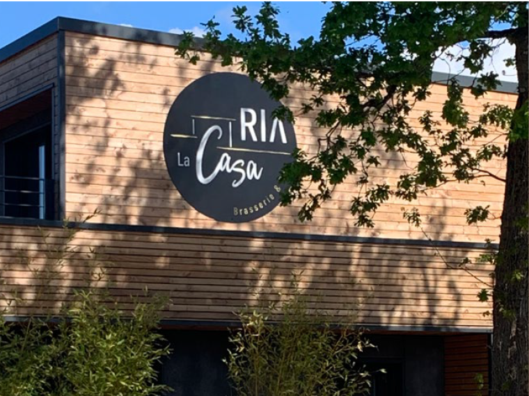 Vue du restaurant La Casa Ria depuis le parking avec l’enseigne au-dessus de la porte et le bâtiment derrière les arbres