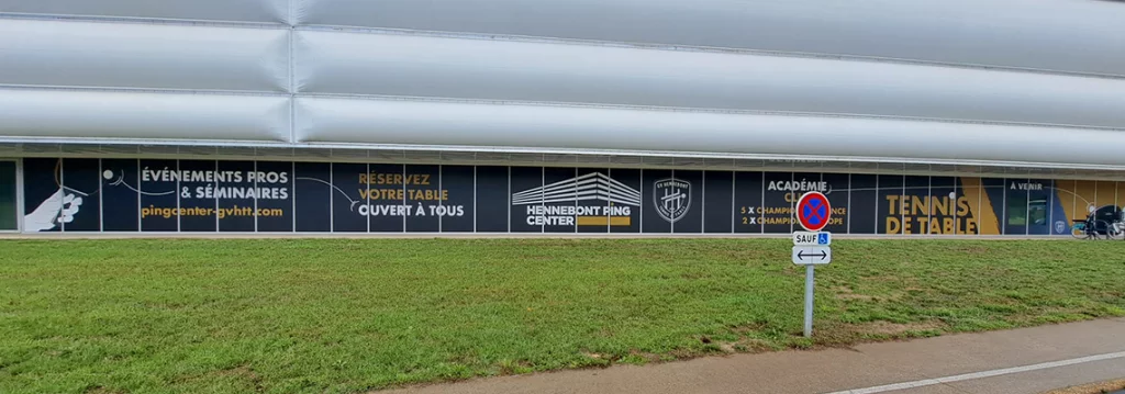 Vue depuis la route de l’habillage des baies vitrées du Hennebont Ping Center, avec illustrations de mains jouant au tennis de table sur fond noir et doré