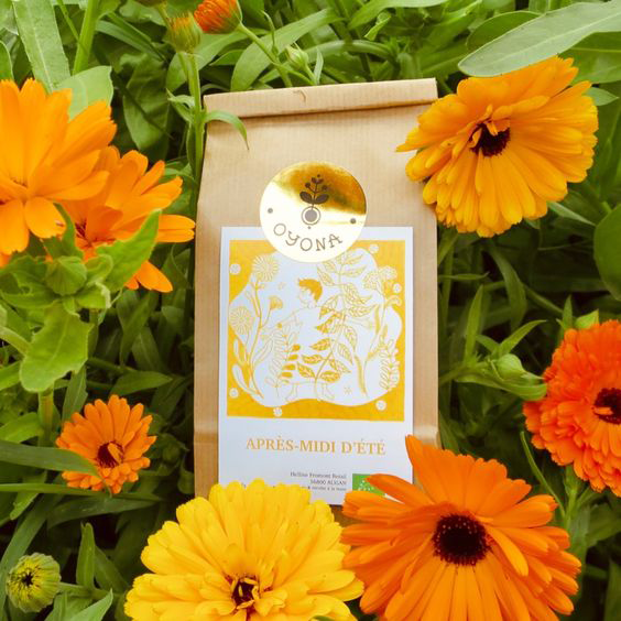 Paquet de tisane « Après-midi d’été » d’Oyona posé au milieu de fleurs orangées, illustration jaune représentant un enfant dans un champ de fleurs